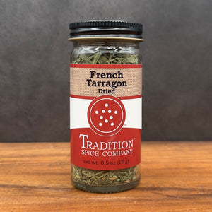A glass jar of French Tarragon from Tradition Spice Company rests on wood, featuring a red and beige label with black text and a central red circle decorated with white dots.