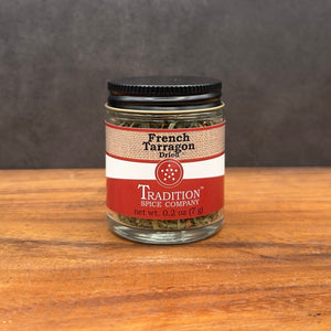 A small glass jar of French Tarragon from Tradition Spice Company, with a red and beige label and black lid, sits on a wooden surface against a dark background—perfect for elevating your dishes.