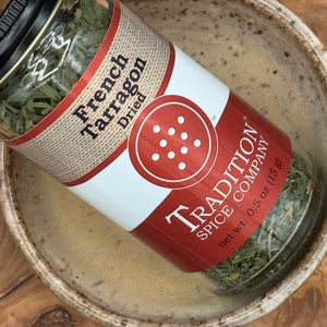 A jar of French Tarragon from Tradition Spice Company sits tilted in a ceramic bowl on a wooden surface. Its red and white label showcases the vibrant green herb, ideal for bringing classic French tarragon flavor to your meals.