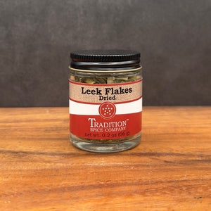 A small glass jar of Leek Flakes sits on a wooden surface with a dark background. The jar features a red and white label, black lid, and contains dried leeks with a mild onion flavor.