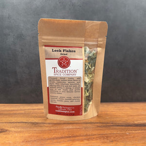 A brown resealable pouch labeled Leek Flakes sits on a wooden surface, featuring a clear window with chopped leeks inside—perfect for adding mild onion flavor to your recipes.