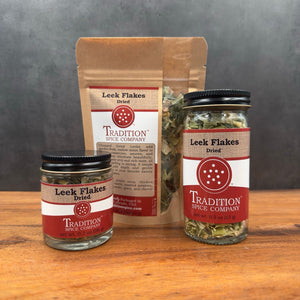 Leek Flakes are shown in a jar, tin, and resealable pouch on a wooden surface against a gray background, highlighting their mild onion flavor.