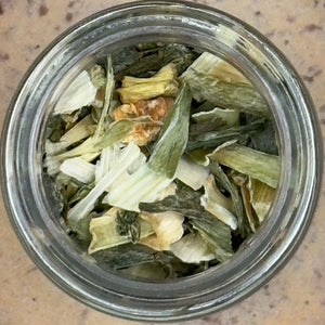 A glass jar of Leek Flakes, prized for their mild onion flavor, is shown from above on a speckled surface.