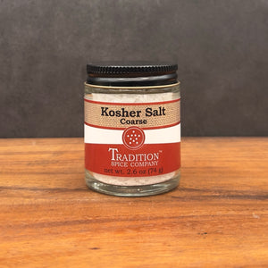 A small glass jar of Coarse Kosher Salt sits on a wooden surface. The jar has a black lid and a red and white label with product details, making it perfect as a cooking salt.