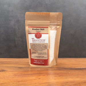 A brown resealable bag labeled Coarse Kosher Salt from Tradition Spice Company sits on a wooden surface against a gray background, featuring red and white branding and essential product details on the front label.
