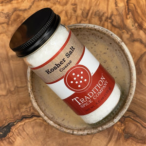 A glass jar of Coarse Kosher Salt rests in a ceramic bowl on a wooden surface. With its black lid and red-and-white label, this salt is ideal for enhancing your favorite dishes with premium seasoning.