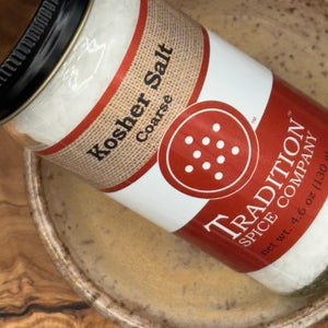 A jar of Coarse Kosher Salt sits by a bowl of light-colored liquid. Its red and tan label with white text makes it ideal for enhancing your favorite recipes as a premium cooking salt.