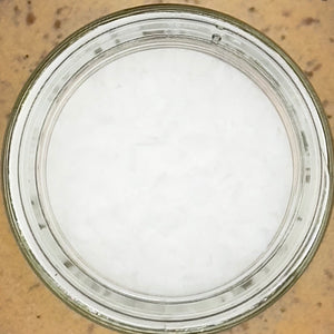 A glass jar of Coarse Kosher Salt flakes, viewed from above, rests on a tan textured surface.