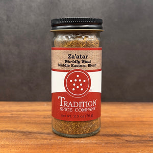 A glass jar of Za’atar, Worldly Wow! Middle Eastern Blend, a classic seasoning with sumac, sits on wood. The jar features a black lid and a red-and-white label with text and a circular logo displaying six white dots.