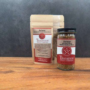 A bag and a jar of Tradition Spice Company's "Za’atar, Worldly Wow! Middle Eastern Blend" with logo and description sit on wood against a dark gray background, showcasing this classic seasoning.