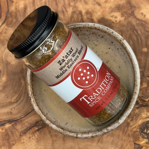 A jar of Za’atar, Worldly Wow! Middle Eastern Blend sits in a ceramic bowl on a wooden surface. The label reads “Za’atar, Worldly Wow! Middle Eastern Blend, net wt. 2.5 oz (70 g).”.