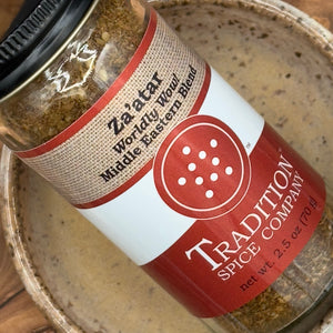 A glass jar of Za’atar, Worldly Wow! Middle Eastern Blend lies on its side on a ceramic dish, displaying the aromatic greenish-brown za’atar mix filled with Middle Eastern seasonings and sumac.