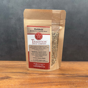 A brown resealable bag labeled "Dukkah, Worldly Wow! Egyptian Blend" sits on a wooden surface with a gray background. A clear window in the bag shows the fragrant Middle Eastern spices inside.