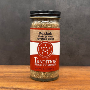 A glass jar of Tradition Spice Company Dukkah Worldly Woul Egyptian Blend spice, featuring a red and white label with a burlap-textured band, set on a wooden surface against a dark gray background.