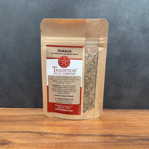 A stand-up pouch of Tradition Spice Company Dukkah spice blend, filled with visible coarse brown and green seeds, set on a wooden surface against a dark gray textured background.