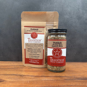 A jar and a stand-up pouch of Tradition Spice Company Dukkah spice blend displayed on a wooden surface against a dark gray backdrop.