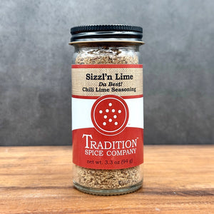 A glass jar of Sizzl’n Lime, Da Best! Chili Lime Seasoning sits on a wooden surface with a gray background. The jar, filled with zesty chili lime spice, has a black lid and a bold red and white label.