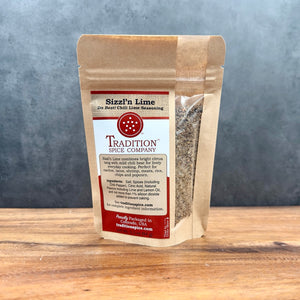 A brown resealable pouch of Sizzl’n Lime, Da Best! Chili Lime Seasoning from Tradition Spice Company rests on a wooden surface with a gray backdrop, displaying the product name and brand logo on its front label.