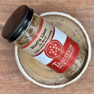 A jar of Sizzl’n Lime, Da Best! Chili Lime Seasoning sits in a shallow ceramic bowl on a wooden surface. The jar features a red label and black lid and holds a zesty chili lime spice blend.