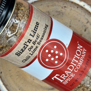 A jar of Sizzl’n Lime, Da Best! Chili Lime Seasoning sits on a tan surface, showing its red and tan label with white text and a circular design, evoking the bold chili lime flavor inside.