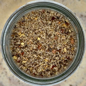 Top view of a glass jar filled with coarse, speckled Sizzl’n Lime, Da Best! Chili Lime Seasoning, featuring grains of salt, pepper, and red pepper flakes. The background is a blurred, neutral-toned surface.