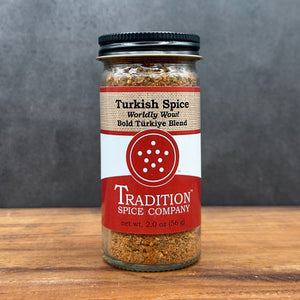 A glass jar of Turkish Spice, Worldly Wow! Bold Türkiye Blend by Tradition Spice Company sits on a wooden surface with a gray background. The label reads Turkish Spice, Worldly Wow! Bold Türkiye Blend and contains 2 oz (56 g).