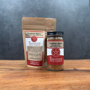 Two containers of Turkish Spice, Worldly Wow! Bold Türkiye Blend—one resealable brown pouch and one clear spice jar with red and white labels—are displayed on a wooden surface against a gray background.