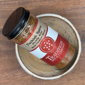 A jar of Turkish Spice, Worldly Wow! Bold Türkiye Blend sits in a small ceramic bowl on a wooden surface. The visible label displays the product name. Ideal as a lamb rub or for elevating your favorite dishes.