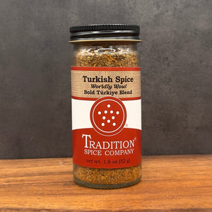 A glass jar of Tradition Spice Company Turkish Spice, featuring a black lid and a red, white, and beige label, set on a wooden surface against a dark gray background.
