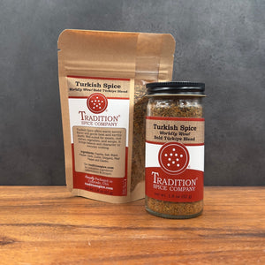 A jar and a stand-up pouch of Tradition Spice Company Turkish Spice, shown on a wooden surface against a dark gray background.