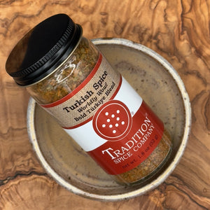 A bottle of Tradition Spice Company Turkish Spice Bold Turkish Blend resting inside a ceramic bowl on a textured wood background.