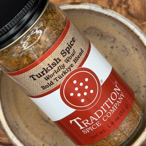 A close-up of a spice jar labeled 'Turkish Spice' by Tradition Spice Company, placed inside a metal bowl. The label is red, white, and beige, and the spice blend inside the clear jar is visible.