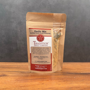 A brown paper pouch labeled "Garlic Mix, Da Best! Garlic Bread Seasoning" from Tradition Spice Company stands on wood, showcasing its salt-free garlic mix and company info, with seasoning visible through a clear side window.