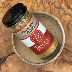 A glass jar of Garlic Mix, Da Best! Garlic Bread Seasoning sits in a small ceramic bowl on a wooden surface. The jar has a red and beige label with a black lid, perfect for elevating your garlic bread recipes.