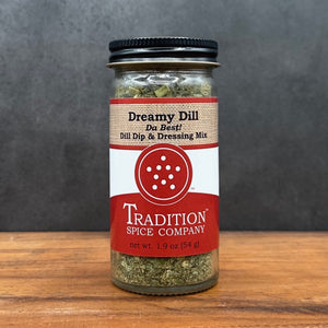 A glass jar of Dream Dill, Da Best! Dill Dip & Dressing Mix rests on a wooden surface, displaying a red and beige label with a circular logo and descriptive text about the blend.