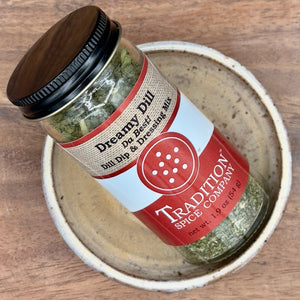 A glass jar labeled Dream Dill, Da Best! Dill Dip & Dressing Mix from Tradition Spice Company rests at an angle in a ceramic bowl on a wooden surface.