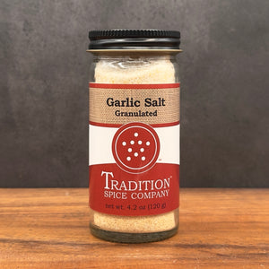 A 4.2 oz glass jar of Granulated Garlic Salt from Tradition Spice Company with a black lid sits on a wooden surface against a dark background.