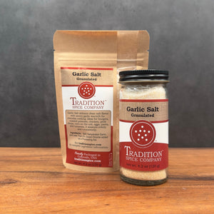 A brown paper bag and a clear glass jar of Granulated Garlic Salt sit side by side on a wood surface against a gray background, both featuring red and white labels.