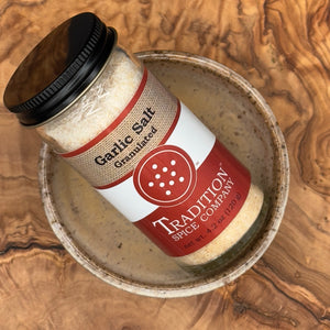 A jar of Granulated Garlic Salt sits at an angle in a speckled ceramic bowl on a wooden surface, ready to enhance any dish with savory flavor.