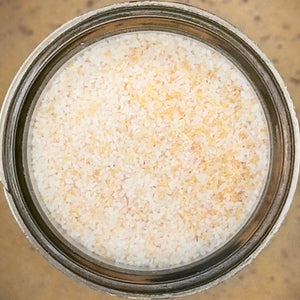 A close-up, top-down view of a glass jar filled with Granulated Garlic Salt from Tradition Spice Company, resembling an off-white seasoning, on a speckled tan surface.