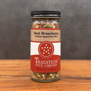 A glass jar of Tradition Spice Company Best Bruschetta Italian Appetizer Mix, featuring a red and white label with a black lid, set on a wooden surface against a dark gray background.