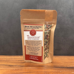 A stand-up kraft paper pouch of Tradition Spice Company Best Bruschetta Italian Spicing Mix, displayed on a wooden surface against a dark gray background.