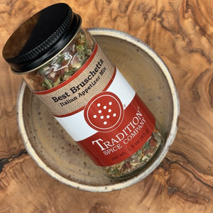 A glass jar of Tradition Spice Company Best Bruschetti Italian Appetizer Mix, featuring a black lid and a red and white label, resting inside a speckled ceramic bowl on a textured wood background.