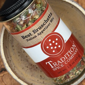 A glass jar of Tradition Spice Company Best Bruschetta Italian Appetizer Mix, featuring a red and white label with a burlap-textured band, resting inside a ceramic bowl.