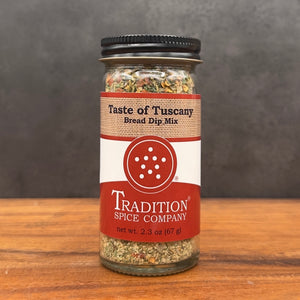 A glass jar of Tradition Spice Company Taste of Tuscany Bread Dip Mix, filled with a dried herb and spice blend, set on a wooden surface against a dark gray background.