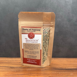 A stand-up kraft paper pouch of Tradition Spice Company 'Taste of Tuscany' bread dry mix, standing on a wooden surface against a dark gray background.