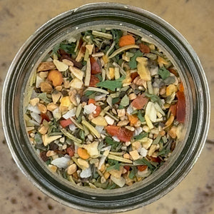 Top-down view of a clear glass jar filled with a colorful dried herb and spice blend containing chopped nuts, seeds, leaves, and small orange pieces, set on a beige textured surface.