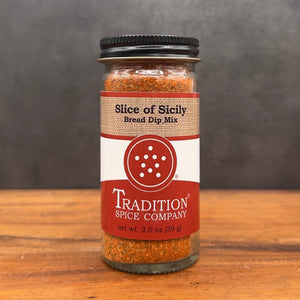 A glass jar of Tradition Spice Company Slice of Sicily Bread Dip Mix with a black lid, set on a wooden surface against a dark gray background.