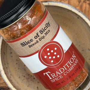 A jar of Tradition Spice Company Slice of Sicily Bread Dip Mix, showing the spice blend through the clear glass container and its label, resting inside a ceramic bowl.