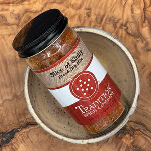 A bottle of Tradition Spice Company Slice of Sicily Bread Dip Mix resting inside a ceramic bowl on a wooden surface.